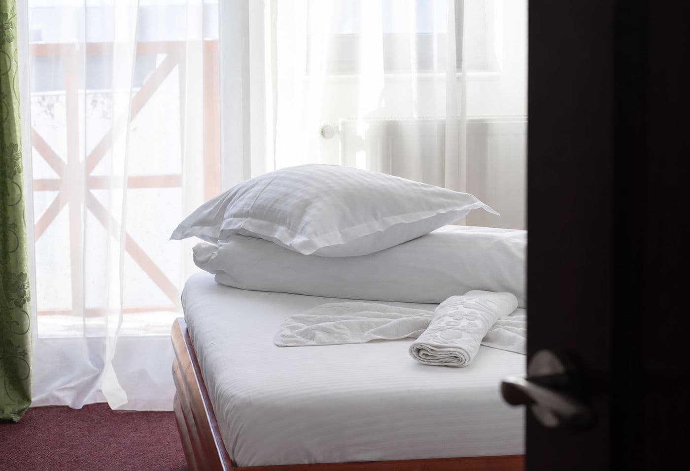 Patul văzut prin ușa întredeschisă, cu balconul din lemn în spatele perdelei